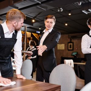 Serious displeased handsome young restaurant manager in formal jacket gesturing hand while giving task to waiter during cleanup
